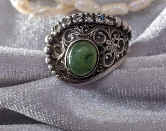 May include: A silver ring with an oval green stone at the center. The ring has intricate floral and scrollwork designs and is accented with small clear stones. A pearl necklace and a gold filigree clasp are in the background.
