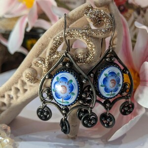 Puede incluir: Pendientes colgantes plateados con diseño floral. Cada pendiente presenta un medallón floral azul y blanco central, rodeado por un marco de filigrana ornamentado y pequeños detalles colgantes. Los pendientes se muestran sobre un fondo suave y texturizado.