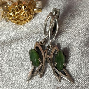 Puede incluir: Pendientes plateados con un diseño en forma de hoja, cada uno con una gema verde vibrante. Los pendientes se muestran sobre una superficie gris texturizada, con un cierre dorado y un collar de perlas en el fondo.