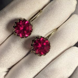 May include: A pair of earrings featuring round, faceted, deep pink stones set in scalloped gold-toned metal settings. Each earring has a curved wire hook for attachment, showcasing a bright, reflective surface on the circular gemstone faces.