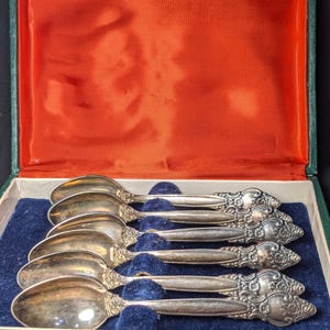May include: A set of six silver teaspoons with ornate handles, displayed in a green velvet-lined box with a red velvet interior.