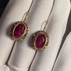 May include: A pair of gold-toned oval earrings with large, faceted, ruby-red gemstones. The earrings have an ornate filigree design around the gemstones and a simple hook closure. The earrings are displayed on a white surface.