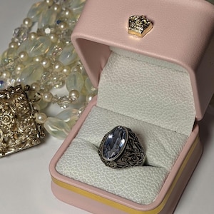May include: A silver ring with a large oval blue stone set in a filigree design. The ring is in a pink ring box with a gold crown on the lid.