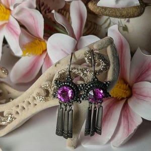 May include: A pair of dangle earrings with a dark gray metal finish. Each earring features a large, round, fuchsia-colored crystal surrounded by a decorative metal design. The earrings have three dangling metal charms.