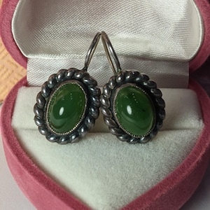 Op de afbeelding: Een paar zilveren oorbellen met groene edelsteen cabochons, gezet in een gedraaid touw ontwerp. De oorbellen zijn in een hartvormige juwelendoos.