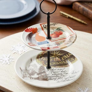 May include: Two-tiered serving stand with a black metal handle. The plates feature vintage-style illustrations of a rooster, tomatoes, and Parisian landmarks. The stand is on a light-colored surface.