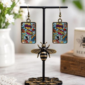 May include: Rectangular drop earrings featuring a colorful comic book pop art print with text bubbles saying WHAM, OOPS, and CRASH. The earrings have bronze-toned metal frames and hooks, displayed on a black metal stand shaped like a bee on a honeycomb.