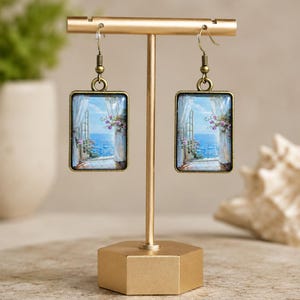 May include: A pair of rectangular earrings with a scenic painting of a window overlooking the sea. The earrings are displayed on a gold-colored stand. The painting features a blue sky, white curtains, and purple flowers.