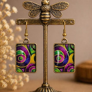 May include: A pair of rectangular earrings with a colorful abstract design in shades of purple, green, and yellow. The earrings are displayed on a decorative brass bee-shaped earring stand. The earrings have a gold-colored frame.