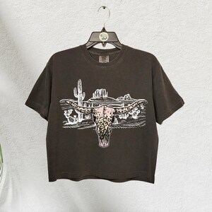 Op de afbeelding: Een bruin T-shirt met korte mouwen met een witte afbeelding van een longhorn-schedel met een roze en bruin luipaardprint. De schedel wordt omringd door een woestijnlandschap met cactussen en bergen.