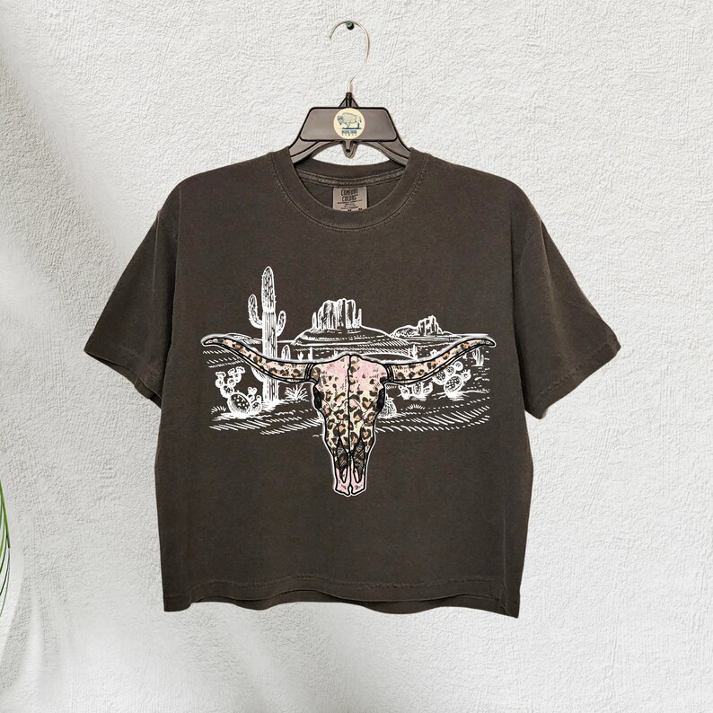 Op de afbeelding: Een bruin T-shirt met korte mouwen met een witte afbeelding van een longhorn-schedel met een roze en bruin luipaardprint. De schedel wordt omringd door een woestijnlandschap met cactussen en bergen.
