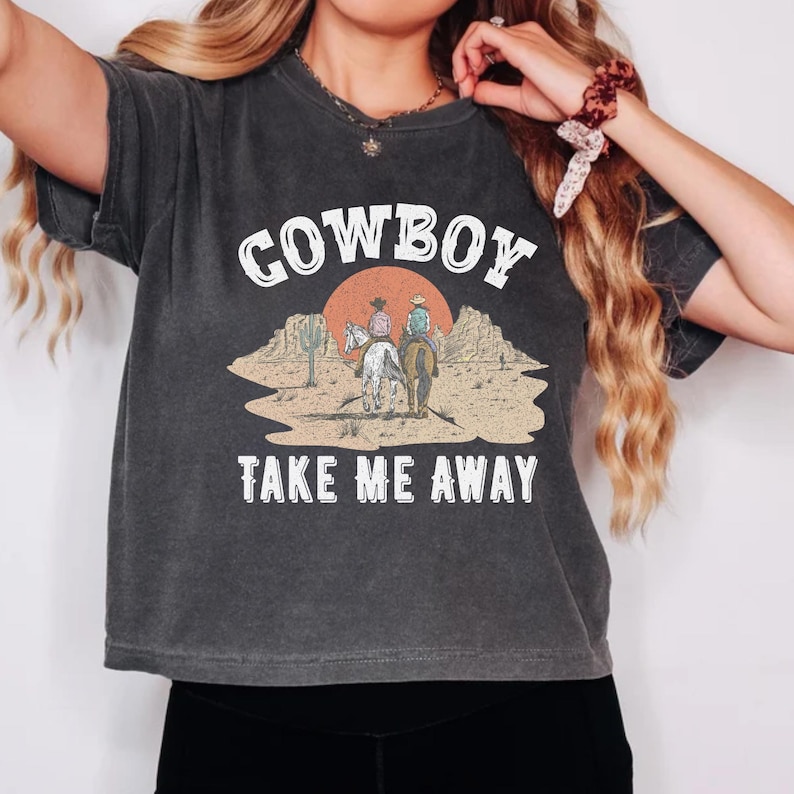 Op de afbeelding: Een donkergrijs T-shirt met een vintage-stijl afbeelding van twee cowboys die op paarden rijden in een woestijnlandschap. De tekst "COWBOY TAKE ME AWAY" is in wit op het shirt gedrukt.