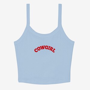 May include: A light blue crop top with thin straps. The word "COWGIRL" is printed in red, arched across the front. The top is a casual style, suitable for warm weather.