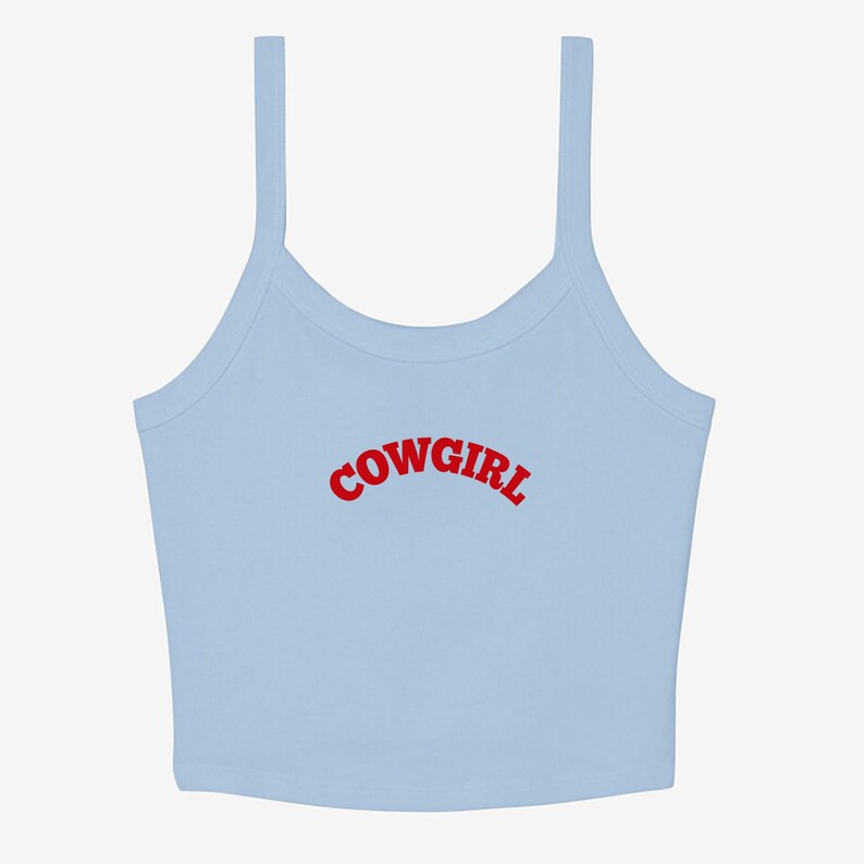 May include: A light blue crop top with thin straps. The word "COWGIRL" is printed in red, arched across the front. The top is a casual style, suitable for warm weather.