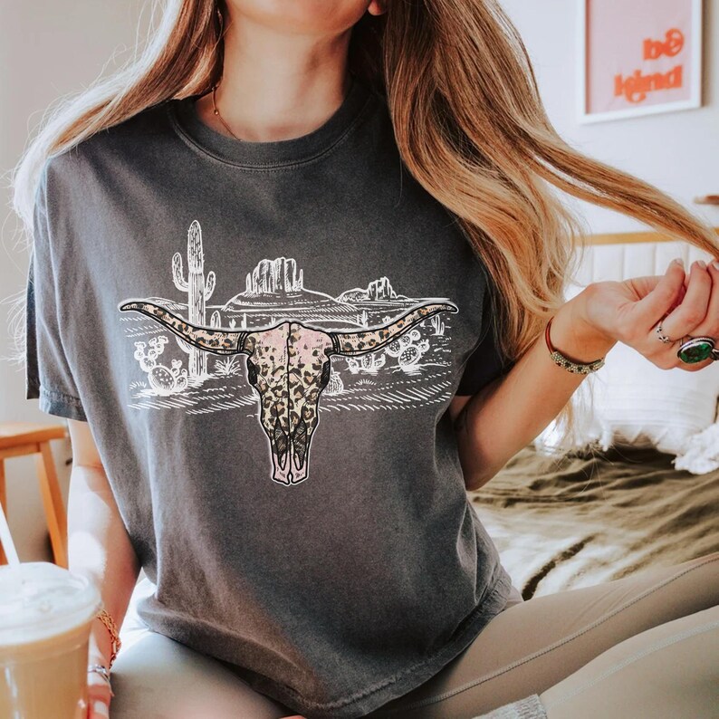 Op de afbeelding: Een donkergrijs T-shirt met een afbeelding van een koeien schedel met een luipaardprint. De schedel wordt omringd door een woestijnlandschap met cactussen en bergen.