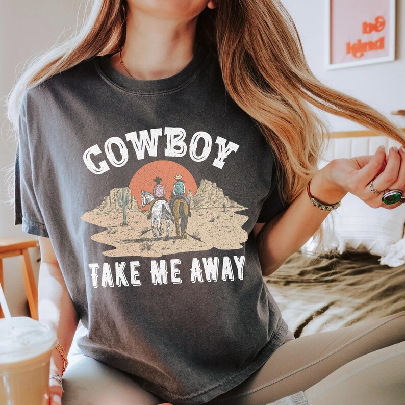 Op de afbeelding: Een donkergrijs T-shirt met een witte grafische print van twee cowboys die op paarden in een woestijnlandschap rijden. De tekst "COWBOY TAKE ME AWAY" is in wit onder de grafische print gedrukt.