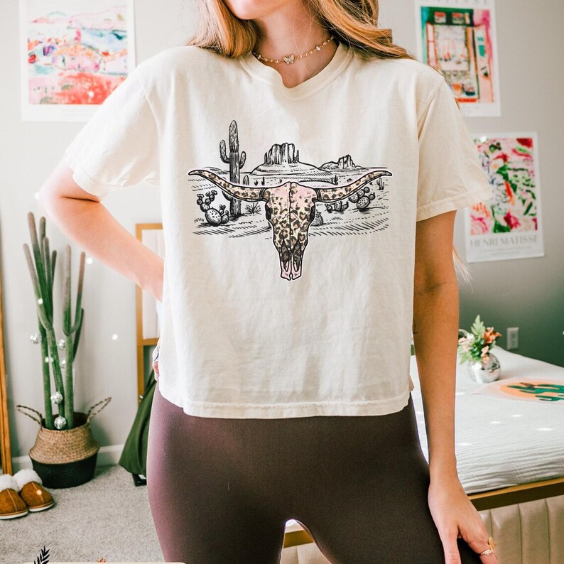Op de afbeelding: Een wit T-shirt met een afbeelding van een luipaardprint koeien schedel met hoorns in een woestijnlandschap. De afbeelding bevat cactussen en bergen.