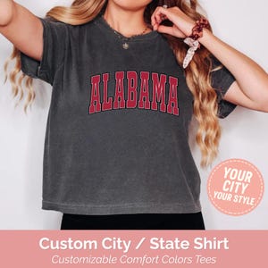 May include: Dark gray t-shirt with the word "ALABAMA" in red block letters. The shirt has a vintage, washed look. A circular graphic with the text "YOUR CITY YOUR STYLE" is visible. The bottom of the image has text that reads "Custom City / State Shirt".