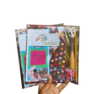 Op de afbeelding: Een knutselset voor het maken van een boek met kleurrijk gestippeld stof, een naald, draad en een potlood. De set bevat instructies voor "Saddle Stitch Bind-A-Book Kit" en "What's Your Story".