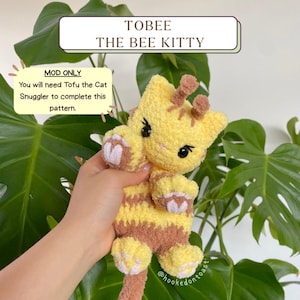 Può includere: Un giocattolo a uncinetto a forma di gattino ape con pelliccia gialla e marrone, che tiene un oggetto a forma di cuore nella sua zampa. Il giocattolo è tenuto da una mano, e lo sfondo è una pianta verde.