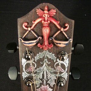 May include: Close-up of a guitar headstock with intricate carvings. The headstock features a stylized figure holding scales, floral designs, and a flower. The guitar has six strings and black tuning pegs.