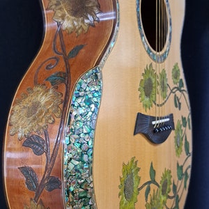 Op de afbeelding: Een houten akoestische gitaar met een natuurlijke afwerking. De gitaar heeft een bloemmotief met zonnebloemen en wijnstokken die in het lichaam zijn gegraveerd. De gitaar heeft een parelmoer inleg rond de rand van het lichaam.