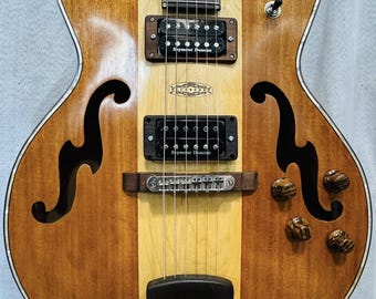 May include: A hollow-body electric guitar with a natural wood finish featuring a light maple center stripe and darker wood sides. It includes two black Seymour Duncan humbucker pickups, two f-holes, a dark wooden tailpiece, and four knobs.