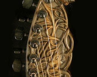 May include: Close-up of the headstock of an acoustic guitar with intricate carvings of a cat and vines. The headstock is made of dark wood and features six tuning pegs. The guitar is labeled "Blueberry".