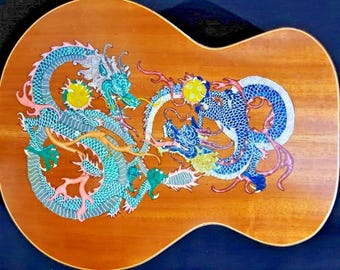 May include: Acoustic guitar back with a detailed dragon design. The dragons are primarily teal and blue, with yellow and pink accents, set against a warm wood grain background. The design is intricate and artistic.