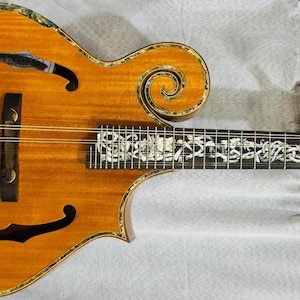 Könnte beinhalten: Eine hölzerne Mandoline mit hellbraunem Finish, mit dekorativen Schnitzereien und einer schneckenförmigen Kopfplatte. Das Griffbrett ist mit aufwendigen Designs eingelegt. Das Instrument hat einen schwarzen Steg und Saitenhalter, mit goldfarbenen Stimmwirbeln.