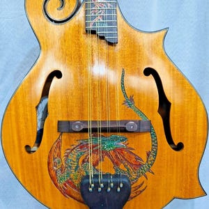 Peut inclure: Mandoline en bois de couleur marron clair avec un motif de dragon. Le corps de l'instrument présente une illustration colorée d'un dragon en rouge, vert et orange. La touche comporte des incrustations décoratives. La mandoline a des ouïes en forme de f et une volute.