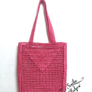 May include: A handmade, pink crochet tote bag with a rectangular shape and a heart-shaped design in the center. The bag has two handles and is made of a textured yarn. The bag is displayed against a white background.