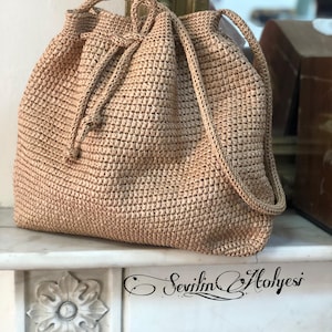 May include: A beige, woven drawstring bag with a rounded shape. The bag features a textured, open weave design and a matching drawstring closure. The bag has a shoulder strap and the text "Sevilin Atölyesi" is visible on the surface.