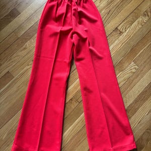May include: Bright red pants with an elastic waistband and flared legs. The pants are made of a smooth fabric and have a classic, retro style. The pants are laid flat on a wooden surface.