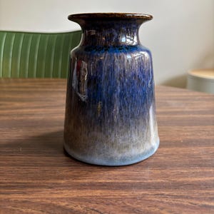 May include: A ceramic vase with a flared rim and a wide base. The vase has a gradient of colors, transitioning from a dark brown rim to shades of blue and gray. The vase is approximately 8 inches tall.