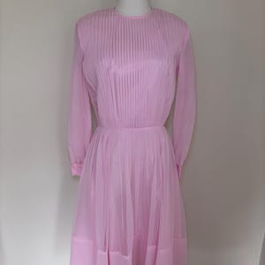 Può includere: Un abito vintage rosa chiaro con maniche lunghe e corpetto plissettato. L'abito ha uno scollo tondo e una gonna fluida. L'abito è esposto su un manichino.