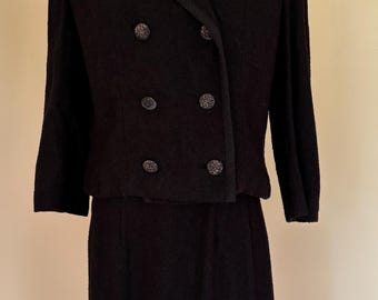 May include: A vintage black skirt suit displayed on a mannequin. The jacket features a double-breasted design with decorative buttons and a contrasting trim. The skirt is a straight, knee-length style. The suit is made of a textured fabric.