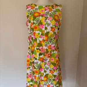 May include: Sleeveless floral print dress in a vibrant mix of colors, including yellow, orange, pink, and green, set against a white background. The dress features a round neckline and a knee-length hemline. The dress is displayed on a mannequin.