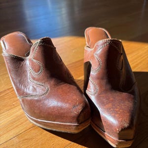 May include: A pair of brown leather clogs with a Western-inspired design. The clogs feature decorative stitching and a wooden sole. The shoes are set on a wooden floor, with sunlight illuminating the details of the footwear.
