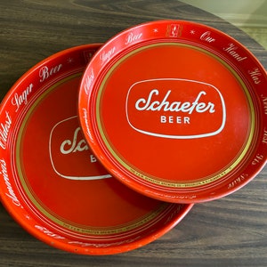 May include: Two vintage red Schaefer Beer serving trays. The trays have a white and gold border with the words "America's Oldest Lager Beer" and "Our Hand Has Never Lost Its Shire". The Schaefer Beer logo is in white.