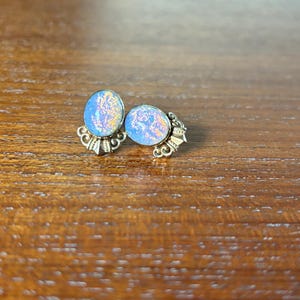 Puede incluir: Un par de pendientes ovalados de color azul iridiscente con engastes de metal dorado. Los pendientes tienen un diseño delicado y ornamentado.