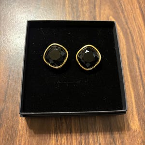 Puede incluir: Un par de pendientes de botón negros, de corte cuadrado, con ribete dorado, presentados en una caja negra. Los pendientes tienen un diseño facetado. La caja está sobre una superficie de madera.