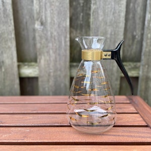 Könnte beinhalten: Eine klare Glaskanne mit schwarzem Griff und goldenen Akzenten. Die Kanne hat eine breite Öffnung und einen schmalen Boden. Sie steht auf einem Holztisch.