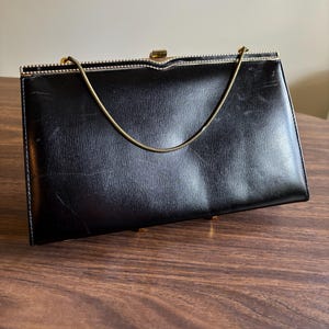 Puede incluir: Un bolso negro vintage con una correa de cadena dorada. El bolso rectangular tiene un cierre de broche y una superficie texturizada. El bolso está sobre una superficie de madera.