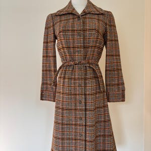 May include: A vintage plaid coat in shades of brown, orange, black, and white, buttoned down the front. The coat features a collared neckline, long sleeves, and a matching belt to cinch the waist. The coat is displayed on a mannequin.