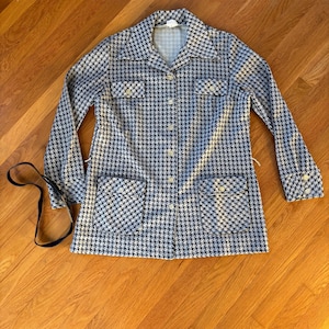 Puede incluir: Chaqueta vintage con estampado de pata de gallo en blanco y negro. La chaqueta tiene mangas largas, cuello y cuatro bolsillos delanteros. Abrochada, con una cinta negra en el lado izquierdo. Se muestra sobre una superficie de madera.