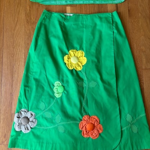 May include: A bright green skirt and top set. The skirt features a wrap design and is adorned with appliqued fabric flowers in yellow, orange, and gray. The top is a button-down shirt with a polka dot pattern.