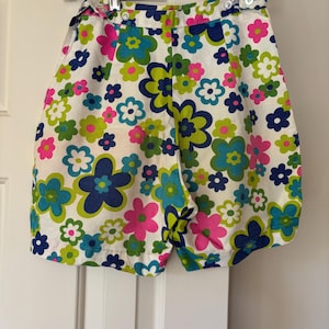 Può includere: Pantaloncini bianchi con un vivace motivo floreale nei toni del blu, rosa e verde. I pantaloncini sono appesi a una gruccia trasparente. I pantaloncini hanno una vita alta e una chiusura con bottoni.