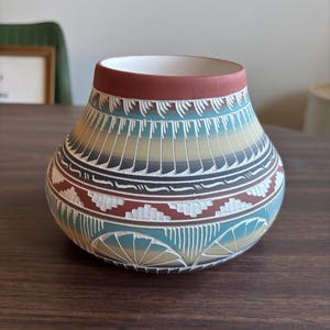May include: A handcrafted ceramic vase with a wide base and a narrow opening. The vase features intricate geometric patterns in shades of turquoise, brown, tan, and white. The rim is a deep red, and the interior is white.