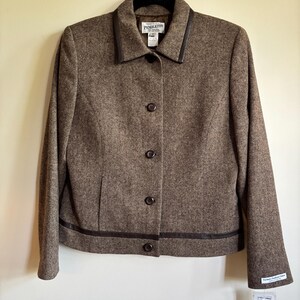 Puede incluir: Una blazer marrón Pendleton con ribete de cuero marrón oscuro en el cuello y el dobladillo. La blazer tiene cuatro botones en la parte delantera y está colgada de una percha negra. La etiqueta dice "Exclusive Pendleton Fabrics".